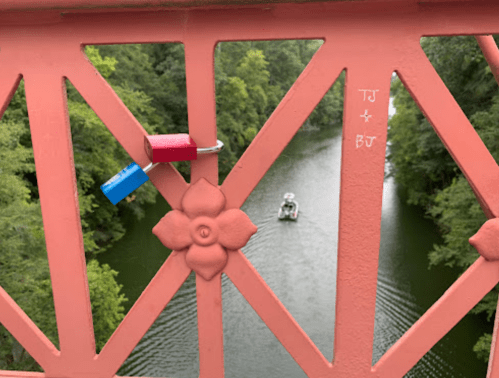 A colorful padlock hangs on a pink bridge, overlooking a river with a small boat and lush greenery in the background.
