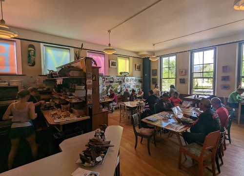 A cozy café interior with patrons seated at tables, a counter with food, and colorful windows letting in natural light.