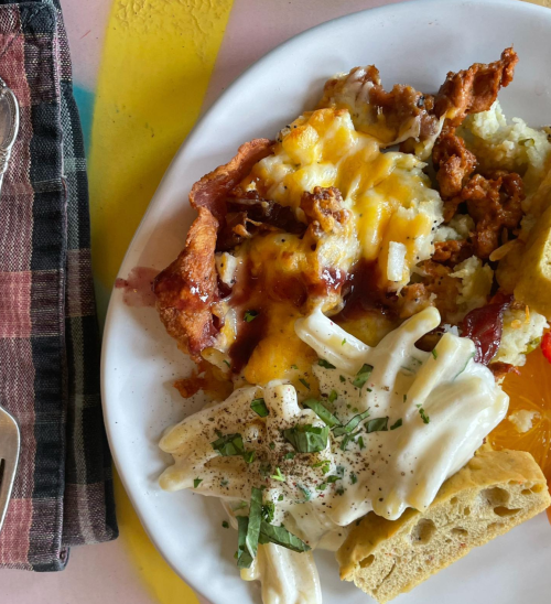 A plate of cheesy casserole topped with barbecue sauce, served with creamy sauce, bread, and colorful fruit.