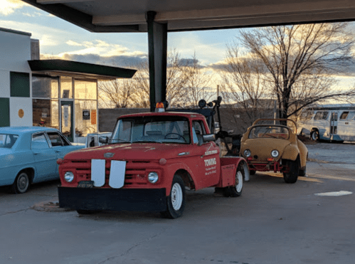 A red tow truck parked at a gas station, towing a yellow vintage car under a cloudy sky.