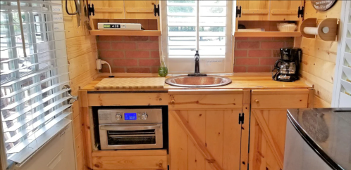 Cozy wooden kitchen with a sink, oven, coffee maker, and open shelves, featuring a brick wall and window.