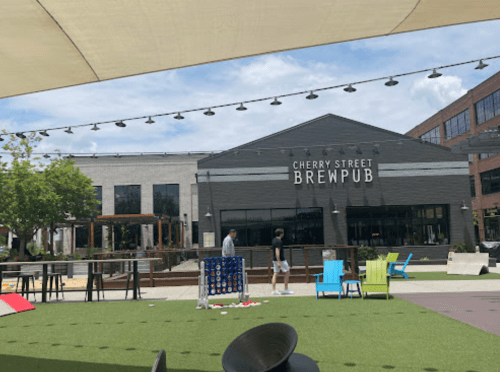 Two people stand outside Cherry Street Brewpub, surrounded by colorful chairs and a green lawn under a sunny sky.