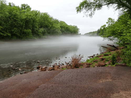 A serene river scene with mist rising over the water, surrounded by lush green trees and rocky banks.