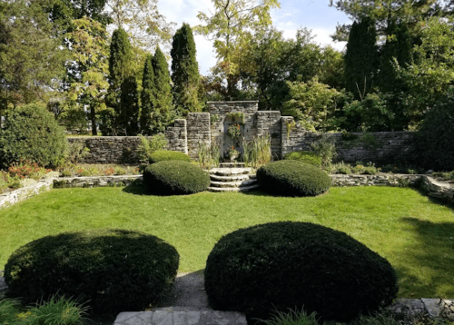 A serene garden featuring manicured bushes, stone walls, and a central fountain surrounded by lush greenery.