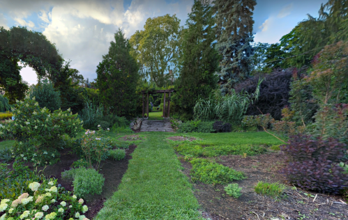 A lush garden path leads to a wooden archway surrounded by greenery and colorful plants under a cloudy sky.