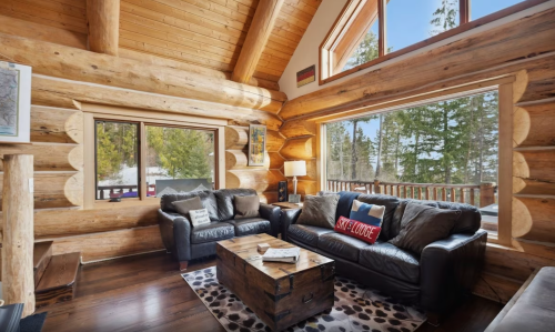 Cozy log cabin living room with large windows, leather sofas, and a wooden coffee table, surrounded by trees.