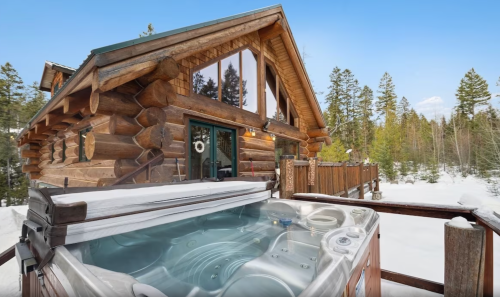 A cozy log cabin surrounded by snow-covered trees, featuring a hot tub on a wooden deck.