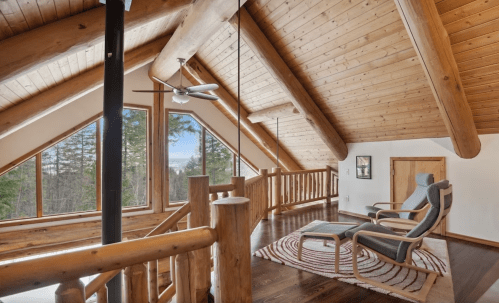 Cozy wooden loft with large windows, a ceiling fan, and a comfortable chair overlooking a forested landscape.