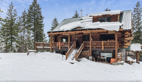 A cozy log cabin covered in snow, surrounded by tall evergreen trees and a clear blue sky.