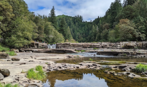 A serene landscape featuring a river, rocky banks, and lush green trees under a partly cloudy sky.