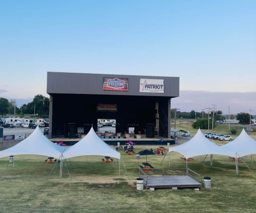 Outdoor stage with a large backdrop, surrounded by tents and seating on a grassy area, under a clear blue sky.