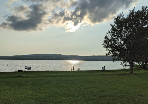 A serene lakeside view with a person standing by the water, reflecting sunlight, and trees in the foreground.