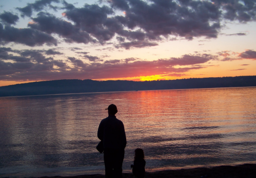 A silhouette of an adult and a child standing by the water, watching a colorful sunset over the horizon.