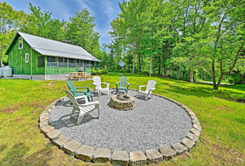 A cozy outdoor seating area with chairs around a fire pit, surrounded by greenery and a green cabin in the background.