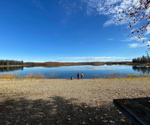 Three people stand by a calm lake under a clear blue sky, surrounded by autumn foliage and distant trees.