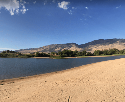 A serene landscape featuring a sandy beach, calm water, and mountains under a clear blue sky.