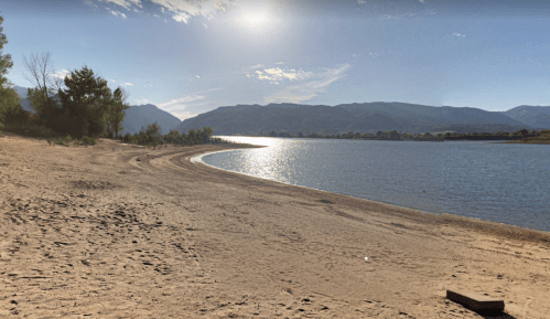 A serene lakeside view with sandy shore, gentle waves, and mountains under a bright sky.