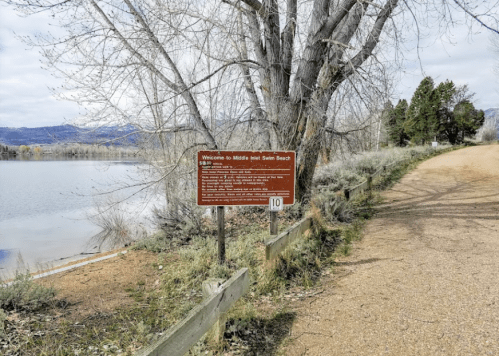 Sign welcoming visitors to Michelle Wells Swim Beach, with a gravel path and trees by a lake in the background.