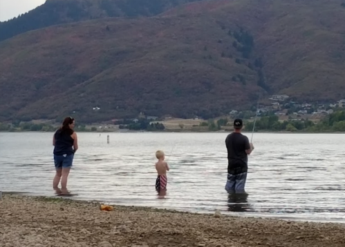 A woman and a man fish in a lake while a child wades in the water, with mountains in the background.