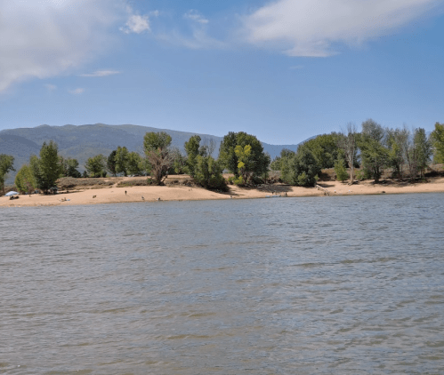 A serene lake scene with sandy shores, green trees, and distant mountains under a blue sky with scattered clouds.