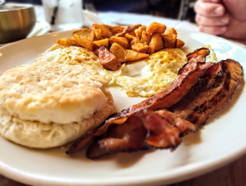 A plate of breakfast featuring scrambled eggs, crispy bacon, seasoned potatoes, and a biscuit.