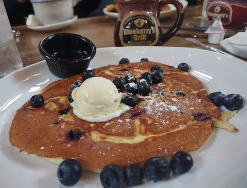 A large pancake topped with blueberries, a scoop of ice cream, and syrup on the side, served on a white plate.