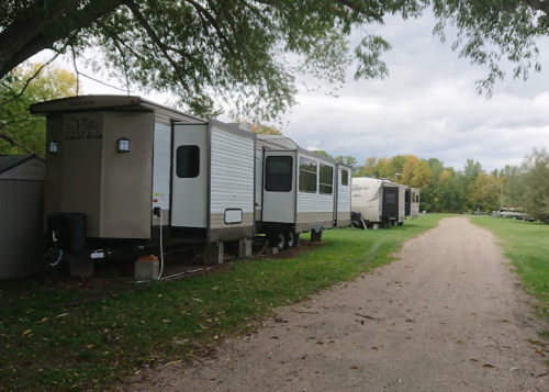 A row of parked trailers along a gravel path, surrounded by trees and greenery under a cloudy sky.