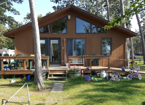 A wooden cabin with large windows, a deck, and a grassy yard, surrounded by trees and outdoor items.