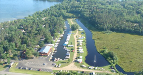 Aerial view of a marina with boats along a winding waterway, surrounded by lush greenery and a parking area.