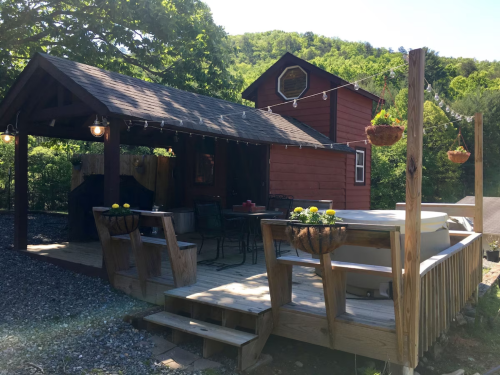 A cozy red cabin with a wooden deck, hot tub, and outdoor seating, surrounded by lush greenery and string lights.