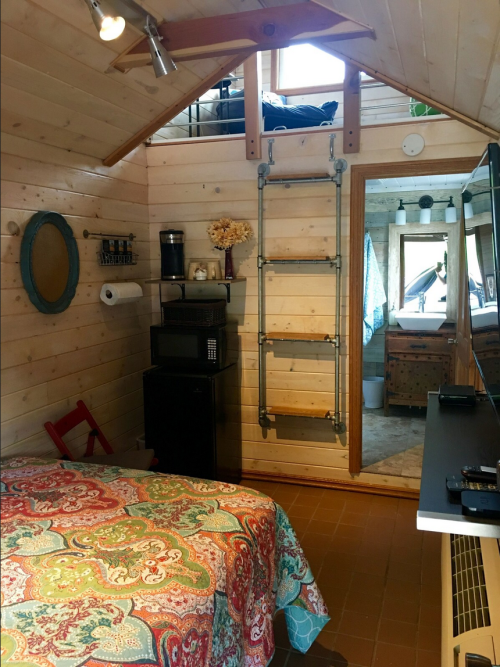 Cozy interior of a tiny house featuring a bed, kitchenette, ladder, and wooden walls. Bright and inviting atmosphere.