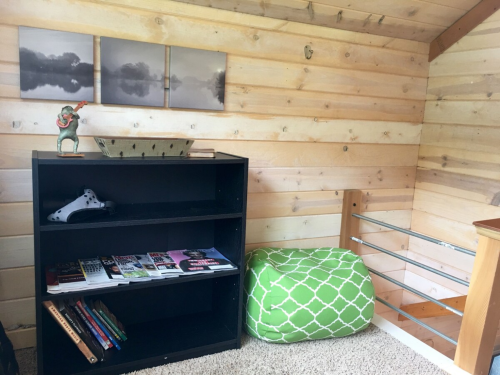 Cozy interior with wooden walls, a black bookshelf, green pouf, and artwork on display.