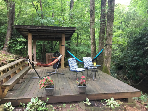 A wooden deck in a forested area with two hammocks, a table, chairs, and potted plants.