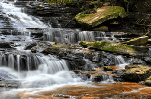 A serene waterfall cascades over smooth rocks, surrounded by lush greenery and gentle flowing water.