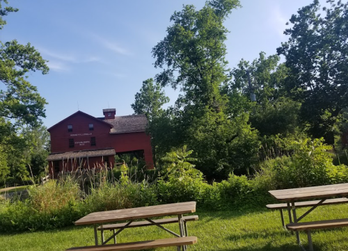 A red building surrounded by greenery and trees, with picnic tables in the foreground under a clear blue sky.