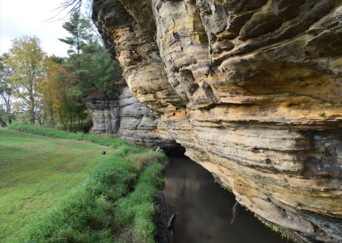 A rocky cliffside with layered stone formations beside a calm, narrow stream and green grass in the foreground.
