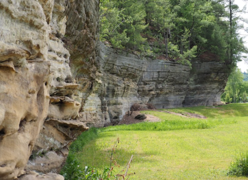A rocky cliffside with layered stone formations beside a grassy area and trees in the background.