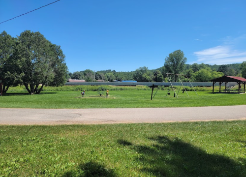 A sunny landscape featuring green fields, trees, a playground, and distant buildings under a clear blue sky.