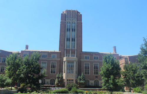 Historic brick building with a tall tower, surrounded by trees and clear blue sky.