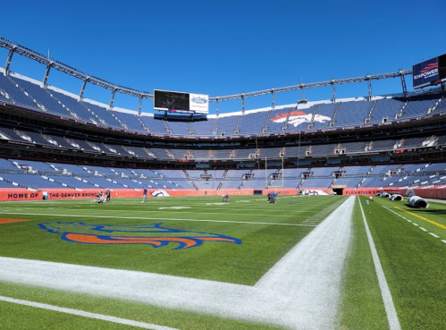 A wide view of a football stadium with blue seats, a green field, and the Denver Broncos logo on the turf.