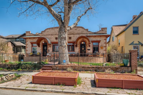 A charming brick house with arched doorways, surrounded by a fence and a large tree, set in a residential neighborhood.