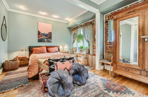 Cozy bedroom with a bed, decorative pillows, a vintage wardrobe, and a large window with plants. Warm, inviting decor.