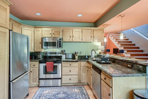 Modern kitchen with wooden cabinets, stainless steel appliances, and a colorful rug, featuring an open layout and stairs.