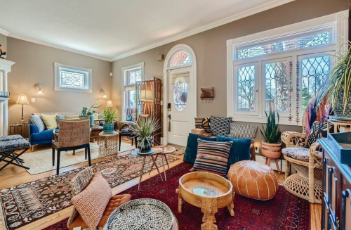 Cozy living room with eclectic decor, plants, and a mix of seating, featuring large windows and warm lighting.