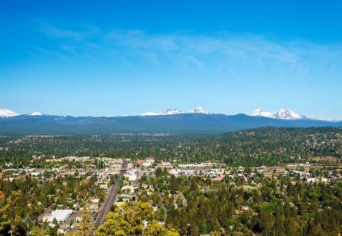 A panoramic view of a town surrounded by lush greenery and mountains under a clear blue sky.