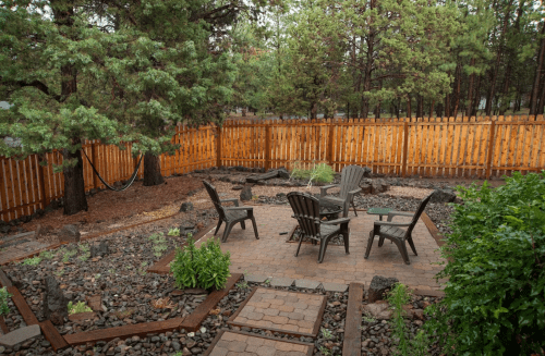 A cozy backyard with a stone patio, four chairs, and a hammock surrounded by trees and a wooden fence.