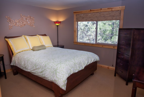 Cozy bedroom with a wooden bed, yellow pillows, a lamp, and a window overlooking trees.