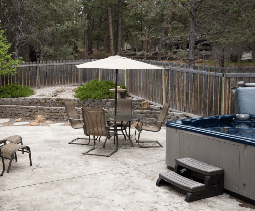 A patio with a hot tub, dining set under an umbrella, and a fenced yard surrounded by trees.