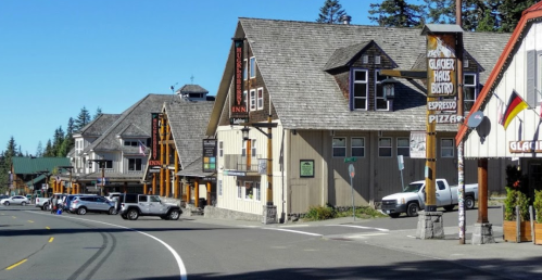 A scenic view of a mountain town with rustic buildings, shops, and parked cars along a quiet road under a clear blue sky.