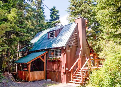 A cozy brown cabin with a green metal roof, surrounded by tall trees and featuring a wooden deck and stairs.